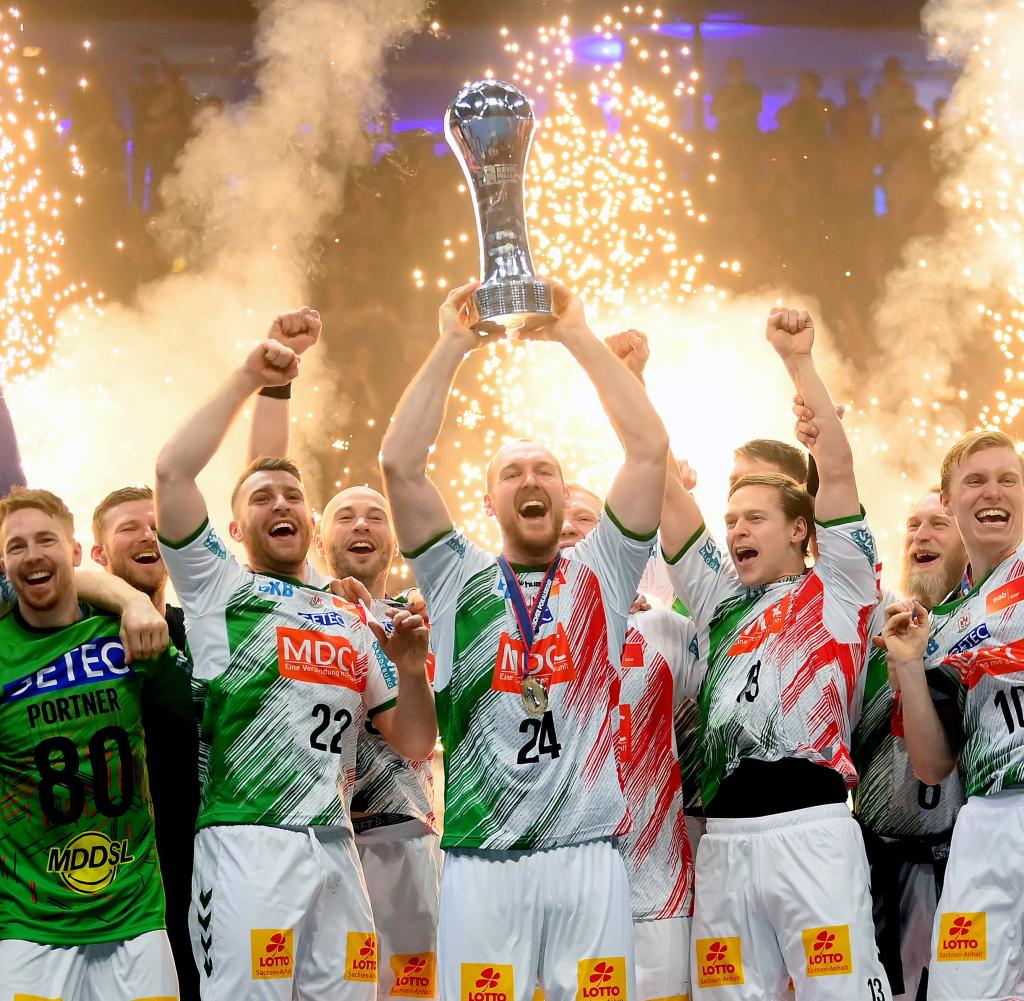 Spieler des SC Magdeburg feiern einen großen Sieg, während sie in ihren rot-grün-weißen Trikots jubeln und ein silbernes Sieger-Trophäe in die Höhe halten; im Hintergrund leuchten goldene Feuerwerksfontänen.