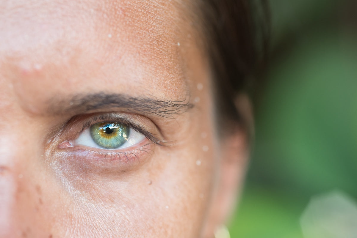 Nahaufnahme eines grünen Auges einer Frau mit leicht gebräunter Haut und Wassertropfen auf dem Gesicht.