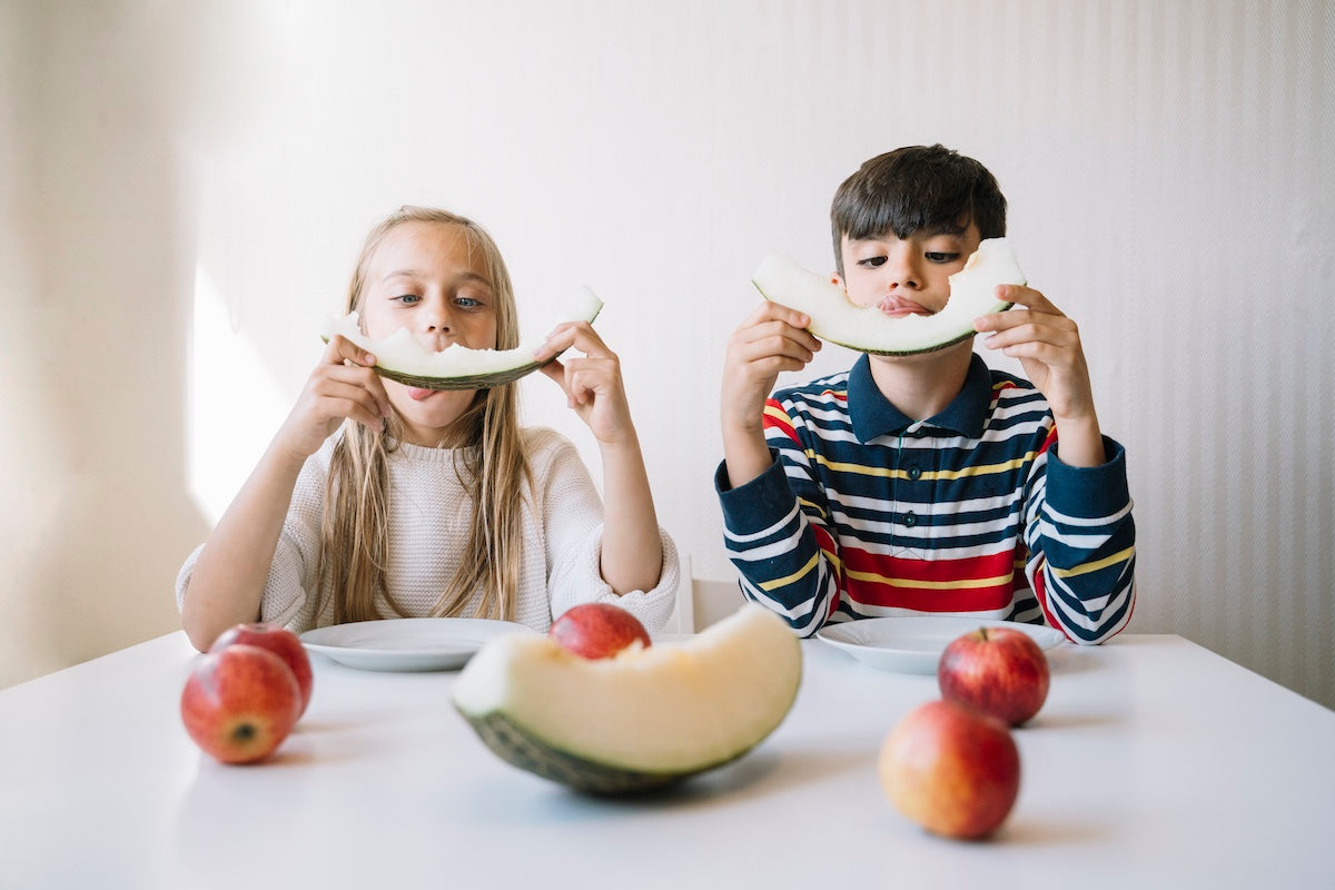 Zwei Grundschulkinder sitzen am Tisch und essen Melonenscheiben mit einem Lächeln im Gesicht, umgeben von frischen Äpfeln – eine spielerische Szene gesunder Ernährung im Kindesalter.