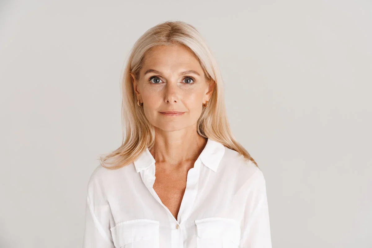 Porträt einer selbstbewussten reifen Frau mit blonden Haaren und weißer Bluse vor neutralem Hintergrund – Symbol für natürliche Schönheit und Pflege.