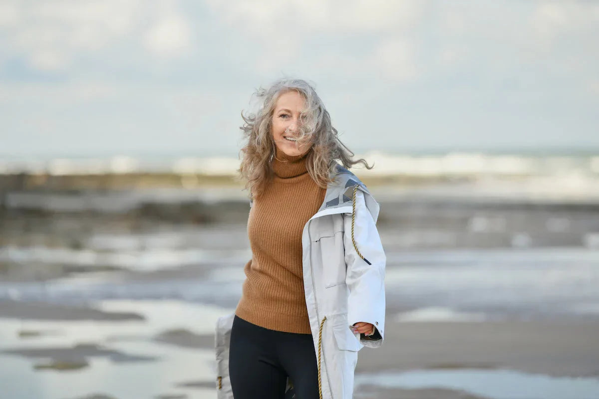 Porträt einer lächelnden Frau mittleren Alters mit grauem Haar, die an einem windigen Strand spaziert.