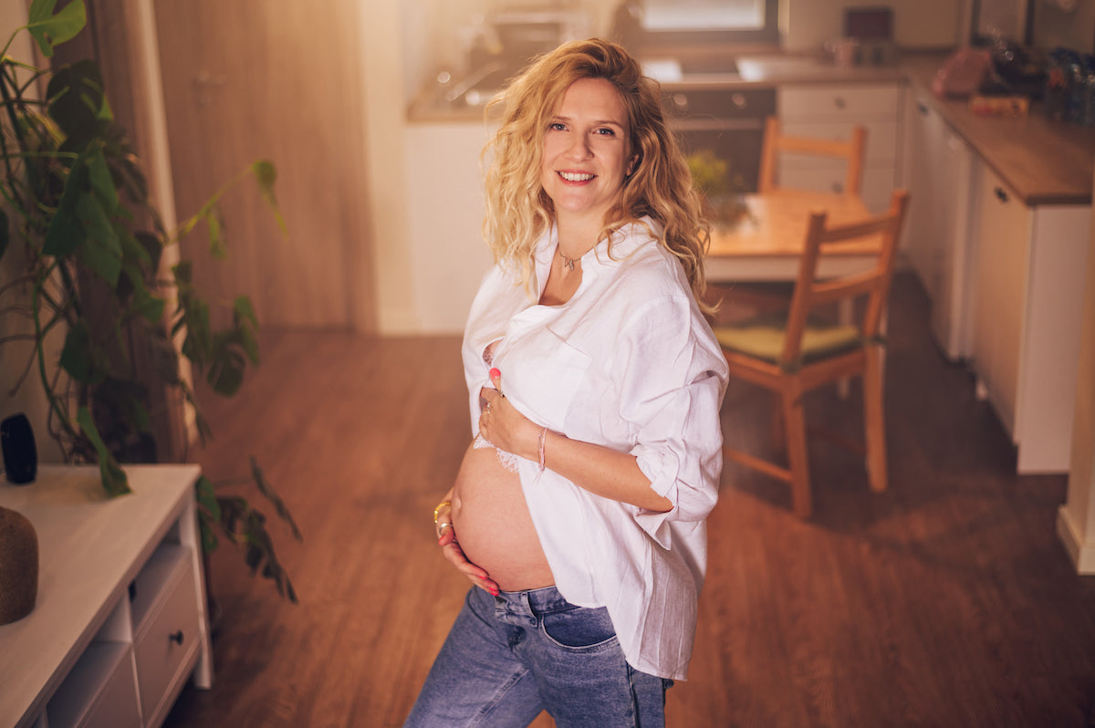 Schwangere Frau mit offenem weißen Hemd steht lächelnd in einer gemütlichen Küche und hält ihre Hand liebevoll auf ihren Babybauch.