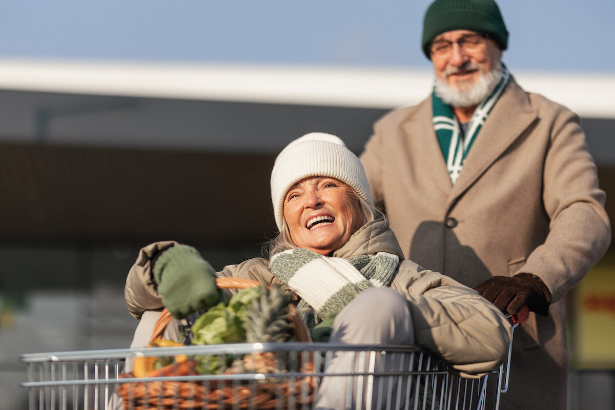Älteres Paar beim Einkaufen im Winter – eine fröhliche Seniorin sitzt im Einkaufswagen mit frischem Obst und Gemüse, während ihr Partner den Wagen schiebt. Beide tragen warme Winterkleidung mit Mütze und Schal.