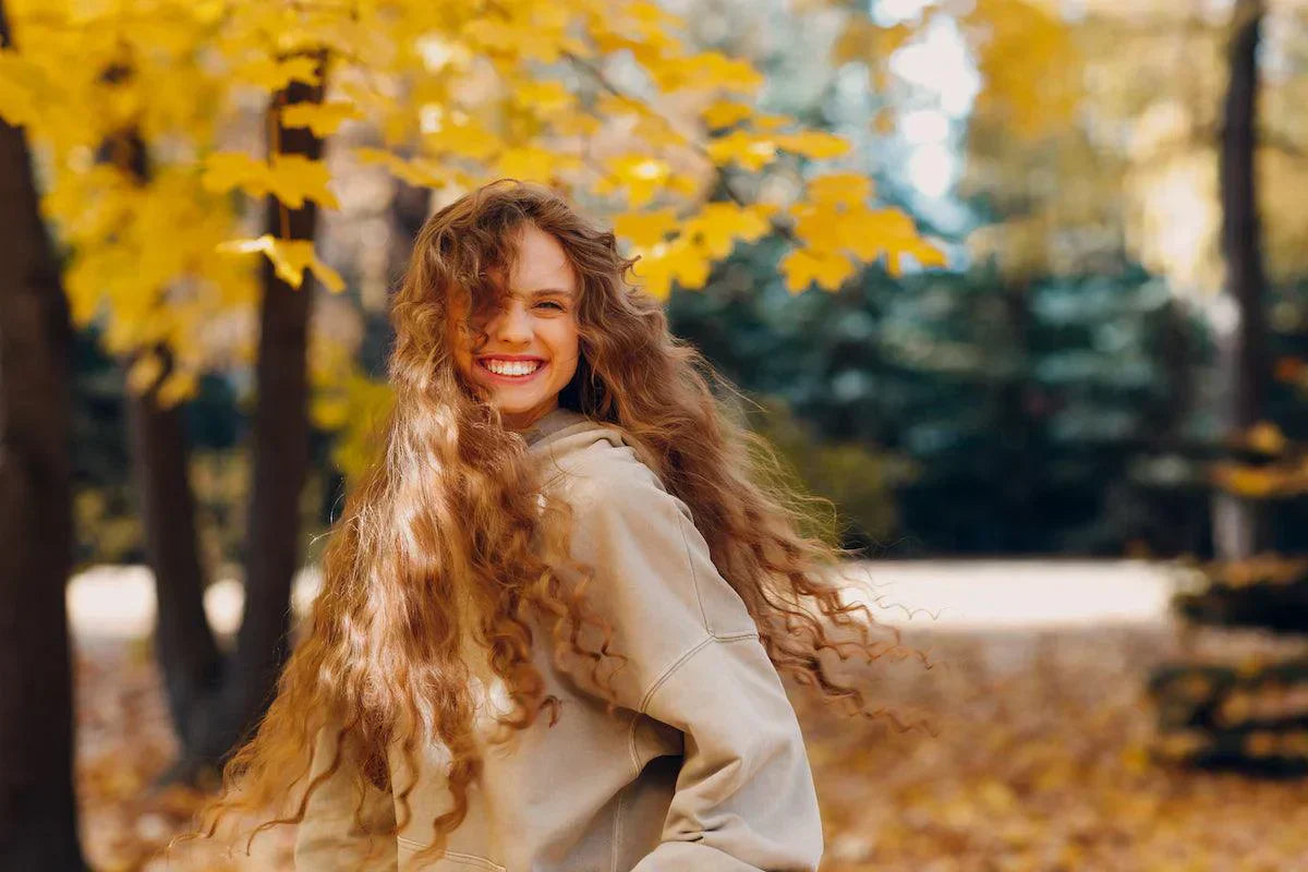 Lächelnde junge Frau mit langen, lockigen Haaren in einem herbstlichen Park, umgeben von gelben Blättern und Bäumen.