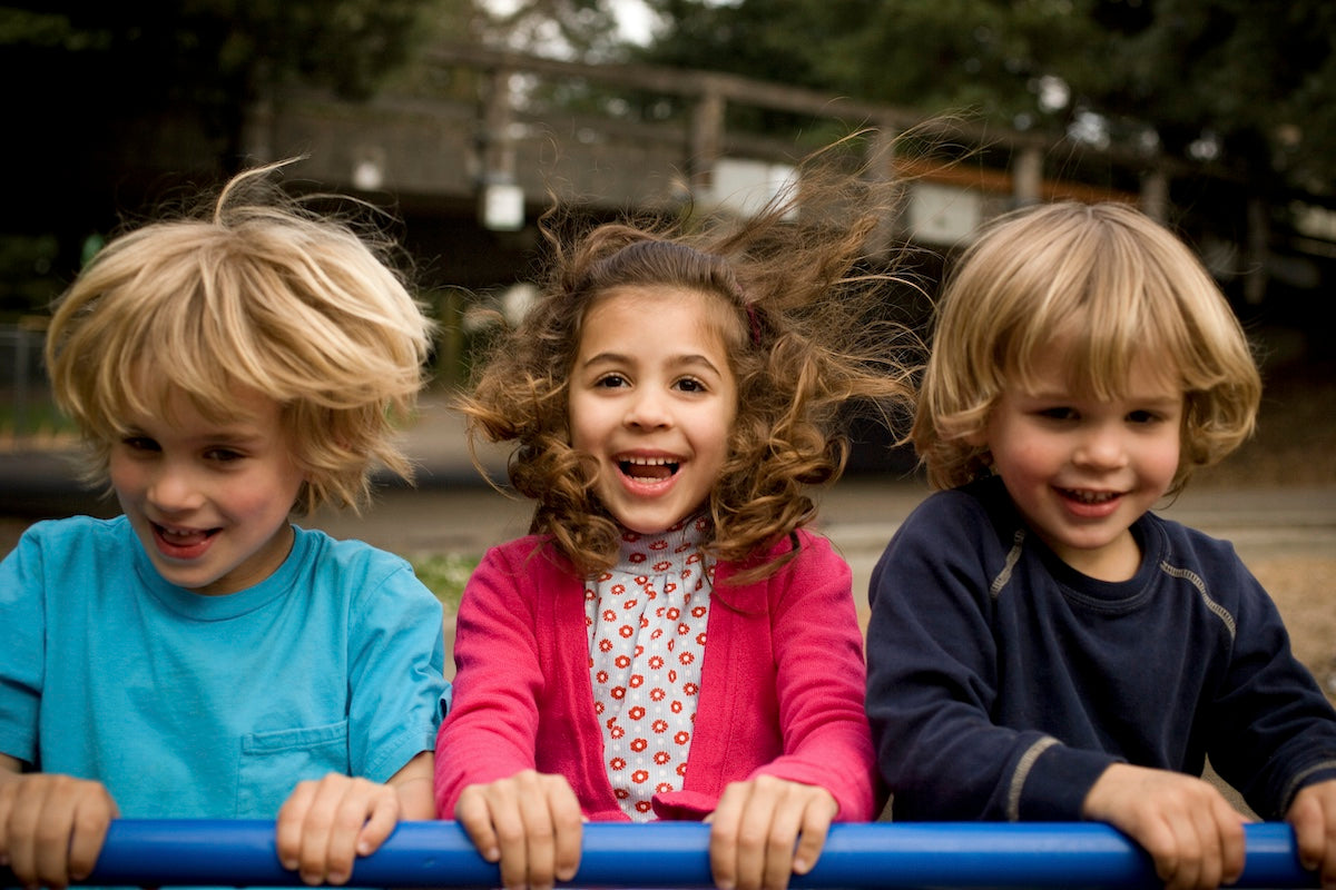 Drei fröhliche Kinder spielen gemeinsam auf einem Spielplatz und lachen in die Kamera.