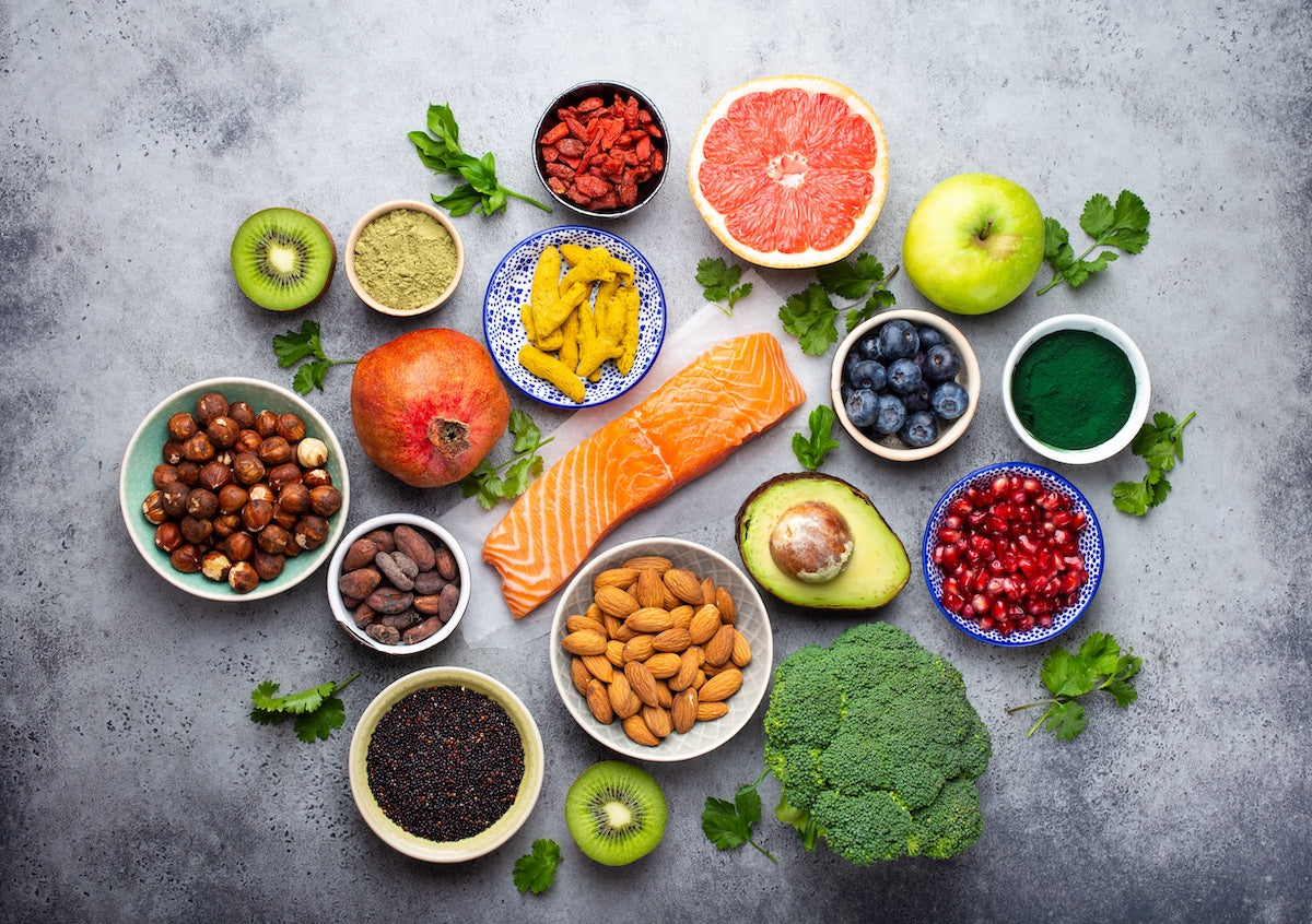 Verschiedene nährstoffreiche Superfoods auf einem Tisch arrangiert, darunter Lachsfilet, Avocado, Brokkoli, Blaubeeren, Granatapfelkerne, Grapefruit, Äpfel, Kiwi, Mandeln, Haselnüsse, Kurkuma, Goji-Beeren, Kakao, Spirulina und schwarzer Quinoa