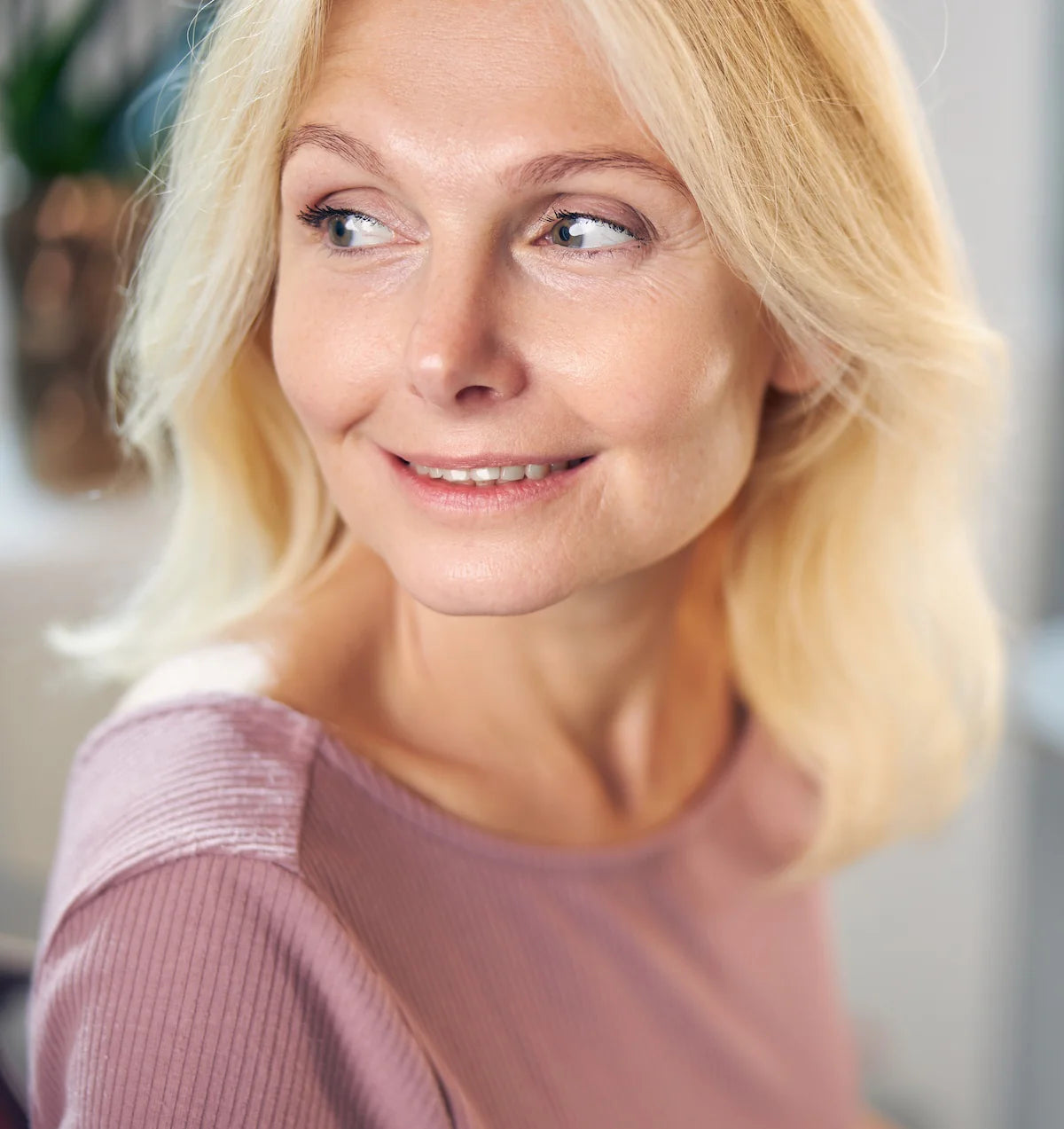 Porträt einer blonden Frau mittleren Alters in einem rosa Oberteil, die seitlich blickt und lächelt, mit einem blauen Punkt auf ihrer Nase markiert.